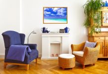 A living room with fireplace mantel featuring blue decorations, blue wingback chair, and wicker chair with matching ottoman.