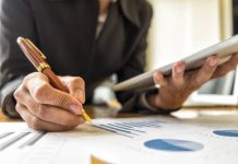 A close-up view shows a person holding a silver tablet in one hand and a pen in the other. Paper with graphs sits on a table.