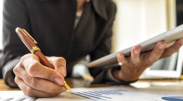 A close-up view shows a person holding a silver tablet in one hand and a pen in the other. Paper with graphs sits on a table.