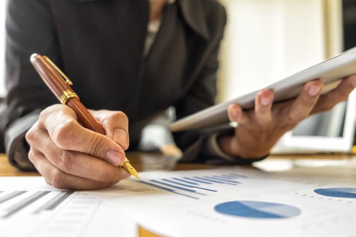 A close-up view shows a person holding a silver tablet in one hand and a pen in the other. Paper with graphs sits on a table.