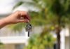 A person's hand dangling a home key in front of a very blurred background of palm trees and a white Caribbean home.