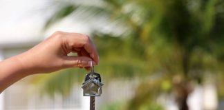 A person's hand dangling a home key in front of a very blurred background of palm trees and a white Caribbean home.