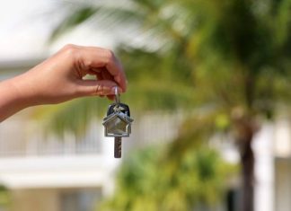 A person's hand dangling a home key in front of a very blurred background of palm trees and a white Caribbean home.