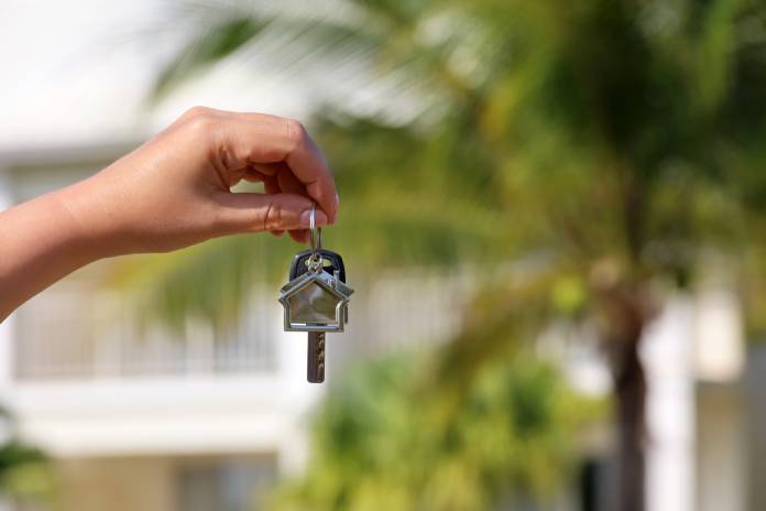 A person's hand dangling a home key in front of a very blurred background of palm trees and a white Caribbean home.