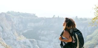 A young woman in shorts and a sleeveless top carries a backpack while looking out over a mountain landscape.