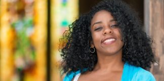 A young Black woman leans against a doorway, smiling happily. Her healthy hair gracefully falls to her shoulders.