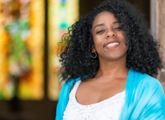 5 Ways To Protect Your Hair From Caribbean Weather on Vacation A young Black woman leans against a doorway, smiling happily. Her healthy hair gracefully falls to her shoulders.