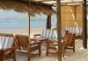 A shaded beachside patio with dried grasses hanging from the roof. The furniture is wooden with blue-and-white cushions.