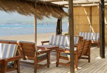 Designing the Perfect Outdoor Lounge for Caribbean Living A shaded beachside patio with dried grasses hanging from the roof. The furniture is wooden with blue-and-white cushions.