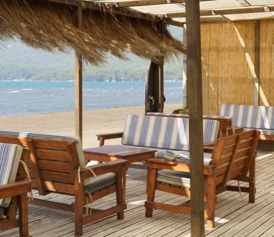 Designing the Perfect Outdoor Lounge for Caribbean Living A shaded beachside patio with dried grasses hanging from the roof. The furniture is wooden with blue-and-white cushions.