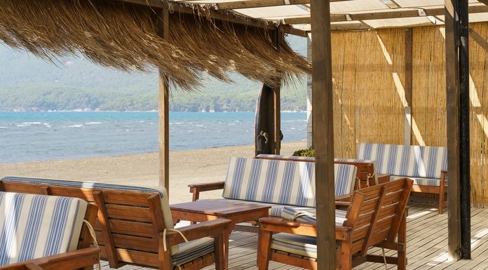 A shaded beachside patio with dried grasses hanging from the roof. The furniture is wooden with blue-and-white cushions.
