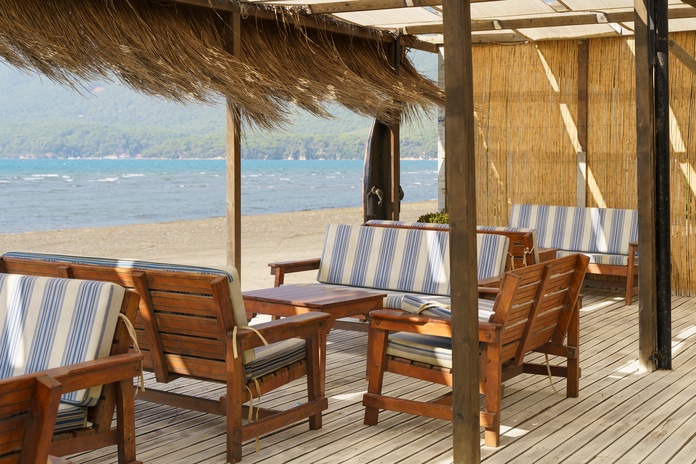 A shaded beachside patio with dried grasses hanging from the roof. The furniture is wooden with blue-and-white cushions.