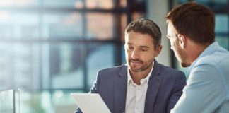 Visual Strategies for Smarter Business Planning A man in a blue suit holds a tablet while sitting next to another man wearing a light blue shirt and holding a pen.