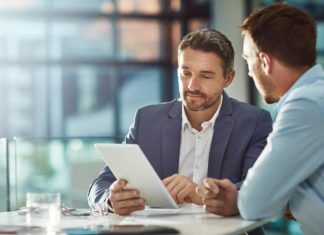 Visual Strategies for Smarter Business Planning A man in a blue suit holds a tablet while sitting next to another man wearing a light blue shirt and holding a pen.