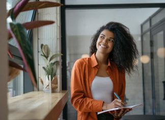 How to Find Your Brand Identity and Market It A person holding a blue pen and a notebook looking out a nearby window. The person smiles while looking outside.