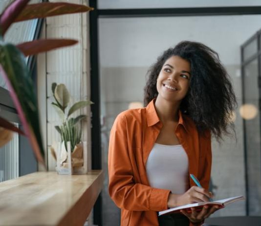 How to Find Your Brand Identity and Market It A person holding a blue pen and a notebook looking out a nearby window. The person smiles while looking outside.
