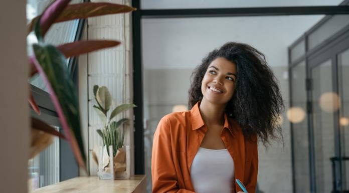 A person holding a blue pen and a notebook looking out a nearby window. The person smiles while looking outside.