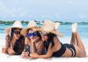 How To Prepare Your Tech for a Beach Vacation Three women in straw hats lie on a sandy beach with blue water in the background. They all look at a phone of them holds.
