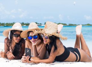 Three women in straw hats lie on a sandy beach with blue water in the background. They all look at a phone of them holds.