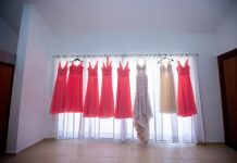 A white gown, six coral gowns, and an ivory gown all hang on a curtain rod as light shines through the sheer curtains.