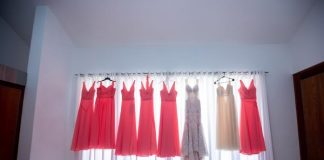 A white gown, six coral gowns, and an ivory gown all hang on a curtain rod as light shines through the sheer curtains.