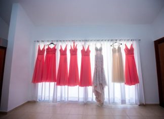 Tips for Picking Stylish Bridesmaid Dresses A white gown, six coral gowns, and an ivory gown all hang on a curtain rod as light shines through the sheer curtains.