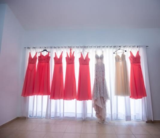 A white gown, six coral gowns, and an ivory gown all hang on a curtain rod as light shines through the sheer curtains.