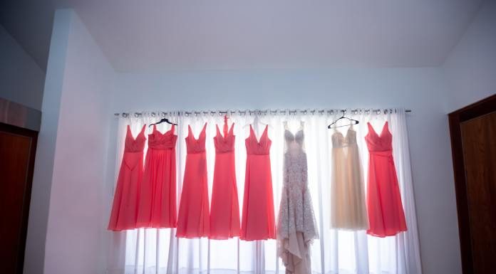 A white gown, six coral gowns, and an ivory gown all hang on a curtain rod as light shines through the sheer curtains.