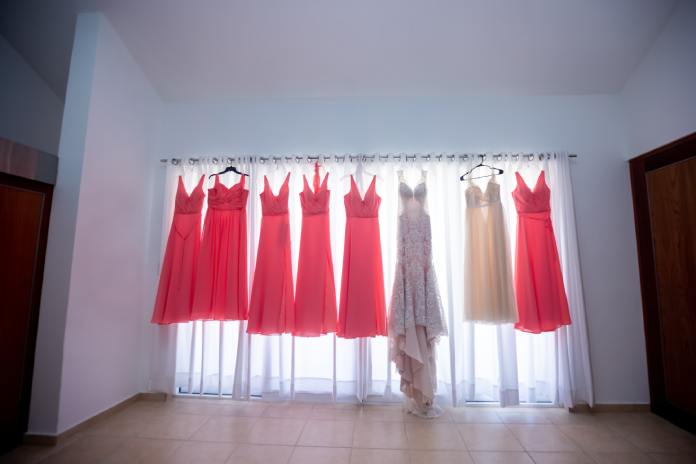 A white gown, six coral gowns, and an ivory gown all hang on a curtain rod as light shines through the sheer curtains.