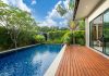 A modern backyard pool beside a wooden deck and glass-walled home, surrounded by lush tropical greenery.