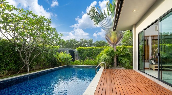 A modern backyard pool beside a wooden deck and glass-walled home, surrounded by lush tropical greenery.