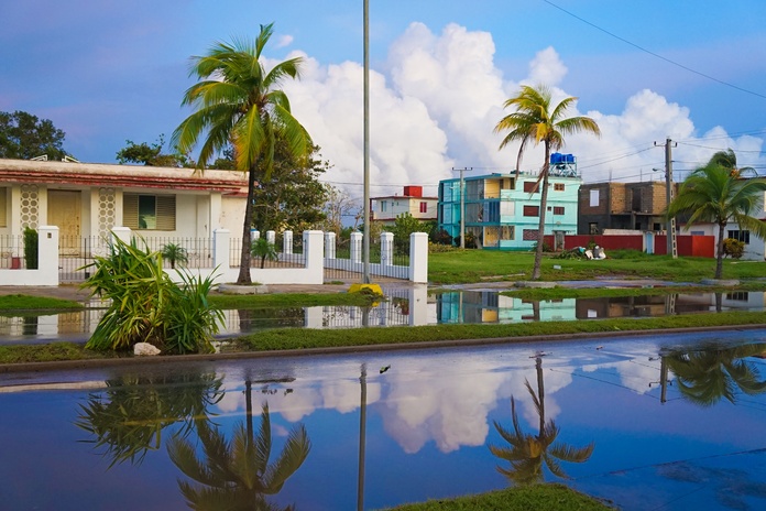 A flooded street in a neighborhood with palm trees and colorful homes reflecting in standing rainwater after a storm. A flooded street in a neighborhood with palm trees and colorful homes reflecting in standing rainwater after a storm.
